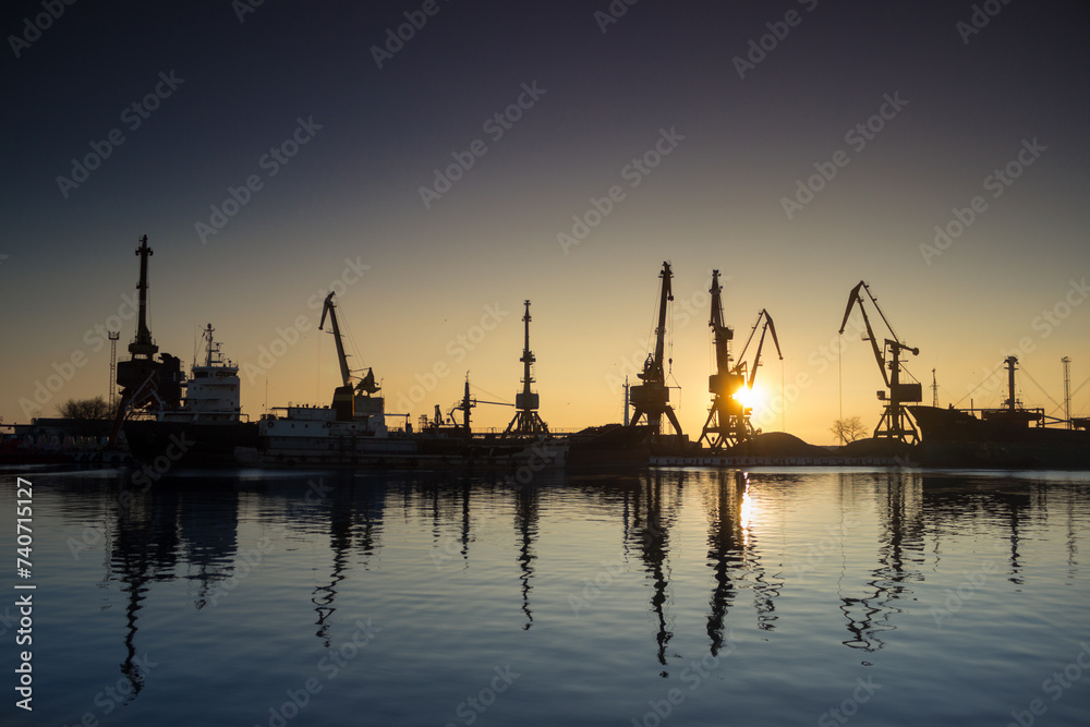 Sunrise silhouettes cargo cranes at busy seaport. Industrial maritime skyline. Trade ships docked, global logistics, export-import operations. Sunrise harbor, reflecting water. Supply chain backdrop.