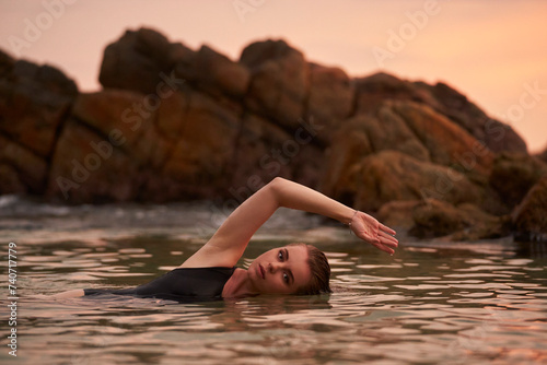 Woman in black swimwear relaxes in sea water at sunset. Female enjoys peaceful ocean swim near rocks. Serene beach bathing  wellness lifestyle. Harmony with nature  leisure activity  relaxation time.