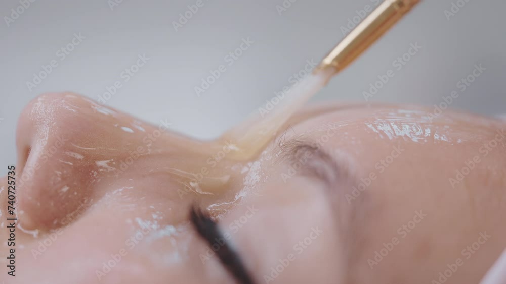 Beautician applies gel face mask on woman's face in beauty clinic, side view.
