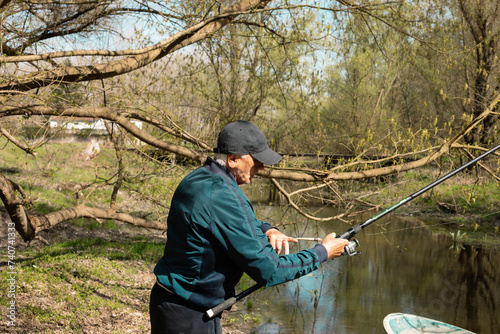 Mature man attaching tackle on rod during fishing. Fisher in casual clothes attaching tackle on rod while standing on riverbank and catching fish from river