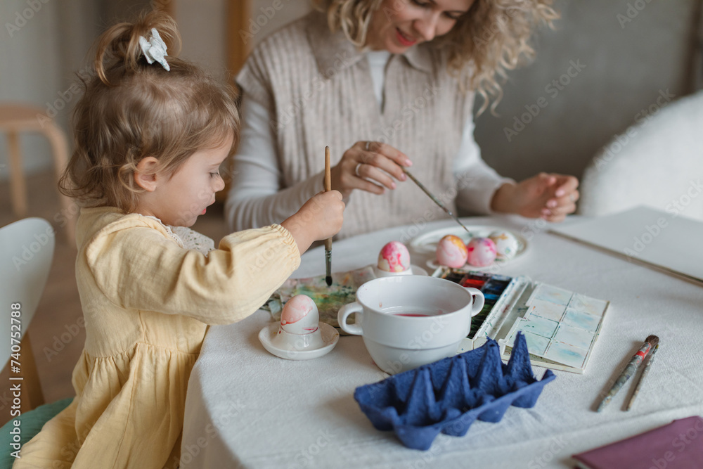 A cute little girl painting eggs with her young stylish mom at a table in a cozy kitchen. Easter family traditions.