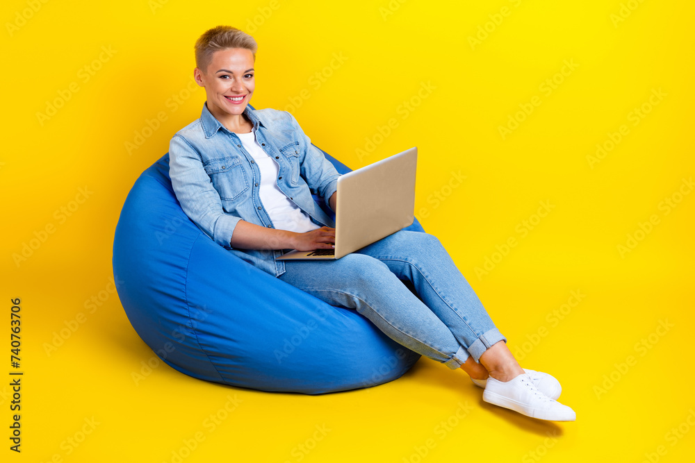 Full body photo of attractive young woman sit bean bag hold netbook dressed stylish denim clothes isolated on yellow color background Full body photo of attractive young woman sit bean bag hold netbook dressed stylish denim clothes isolated on yellow color background