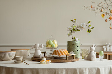 Interior design of easter dining room interior with round table,  colorful eggs, easter cake, easter bunny sculptures, vase with flowers, beige bowl and personal accessories. Home decor. Template.