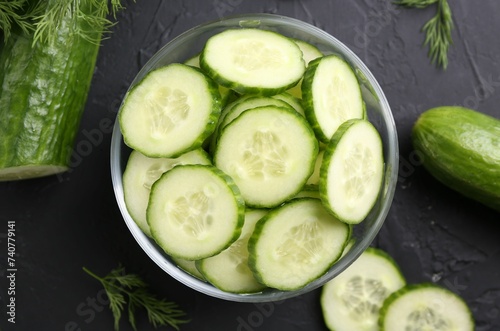 Cut cucumber in glass bowl, fresh vegetables and dill on dark gray textured table, flat lay
