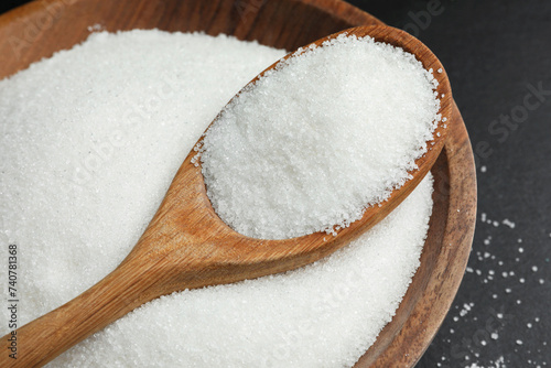 Granulated sugar in bowl and spoon on black table, closeup