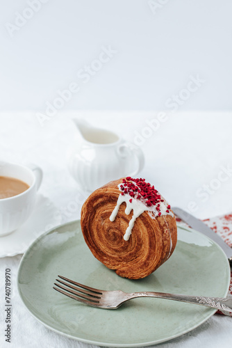 There is a round croissant on a green plate.next to it is a napkin with red patterns and vintage cutlery, a cup of coffee,a milk jug .White background