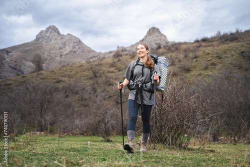 Young carefree female traveler walks with grey backpack and trekking poles outdoors hiking and exploring , smiling tourist is cheerful and happy in the mountains, journey tourism 