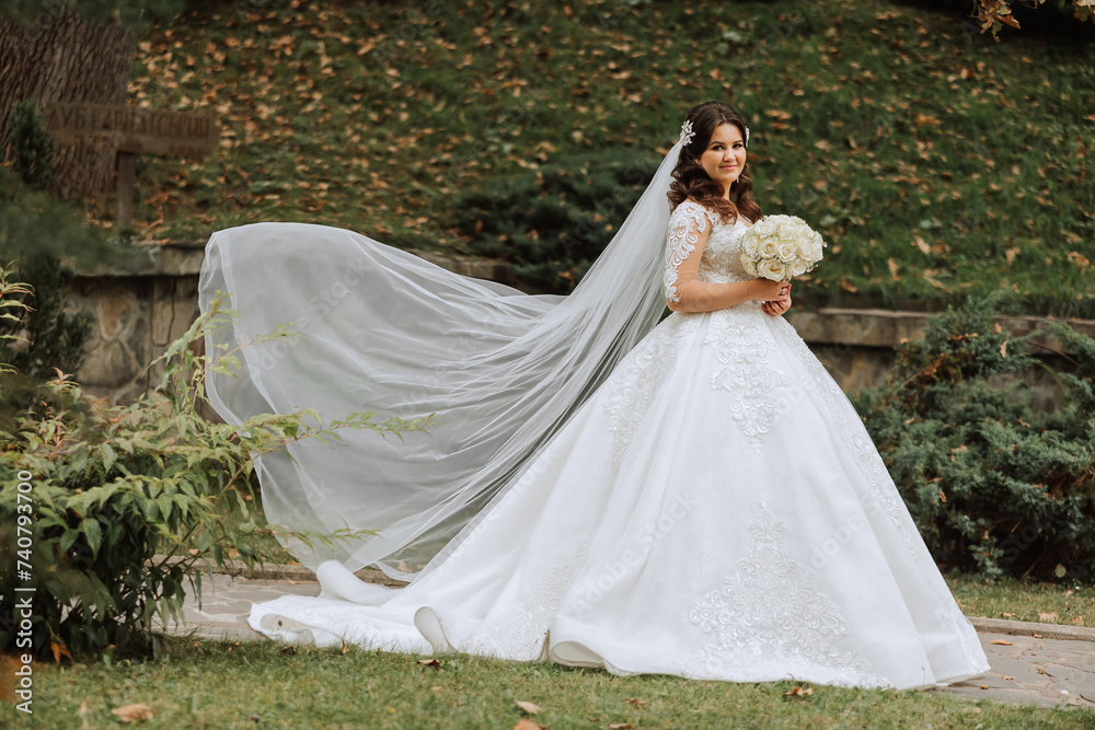 A brunette bride in a voluminous white dress holds a bouquet, poses. The veil is thrown into the air. Beautiful hair and makeup. Wedding photo session in nature.