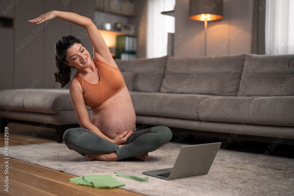 A beautiful, happy, young pregnant woman doing yoga exercises at home and watching a video tutorial on a laptop