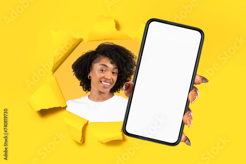 Cheerful young african american woman showing smartphone with empty screen through hole in paper © Prostock-studio