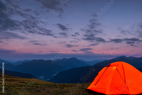 Glowing orange tent in the mountains under dramatic evening sky, mountains in the background. created generative ai