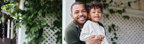 happy african american father in braces hugging cute curly son and sitting on porch on house, banner photo