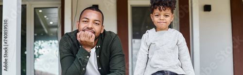 joyful african american father and son smiling and sitting on porch of house, family banner