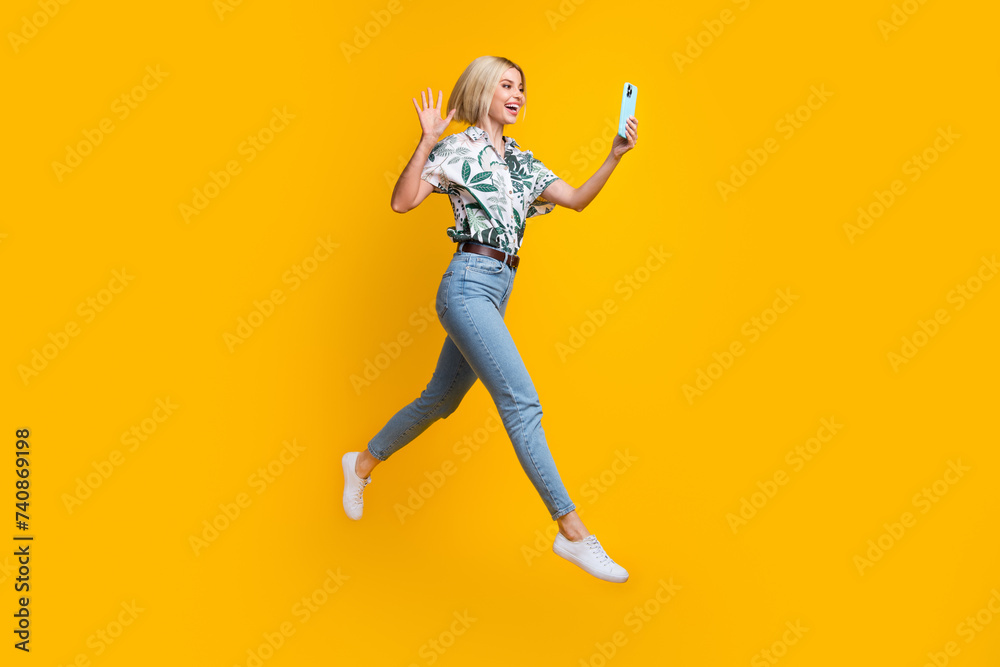 Full size photo of friendly nice girl wear print blouse jeans flying talk by video call on smartphone isolated on yellow color background Full size photo of friendly nice girl wear print blouse jeans flying talk by video call on smartphone isolated on yellow color background