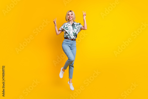 Full length photo of good mood adorable girl dressed print shirt jumping showing v-sign symbol isolated on yellow color background