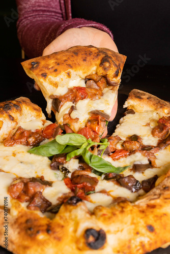 Pizza tradizionale napoletana con mozzarella, melanzane a funghetto fritte, pomodori e basilico fresco servita in una pizzeria e con una fetta tagliata e mantenuta da una cliente