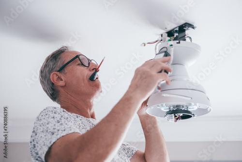 Mature experienced electrician on a ladder installing a paddle fan on the ceiling in the living room at home in preparation for the upcoming summer