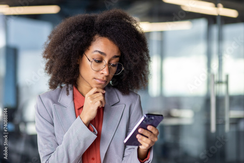 Schilderij op canvas Thoughtful latino woman in grey suit holding chin with hand while looking at smartphone screen indoors