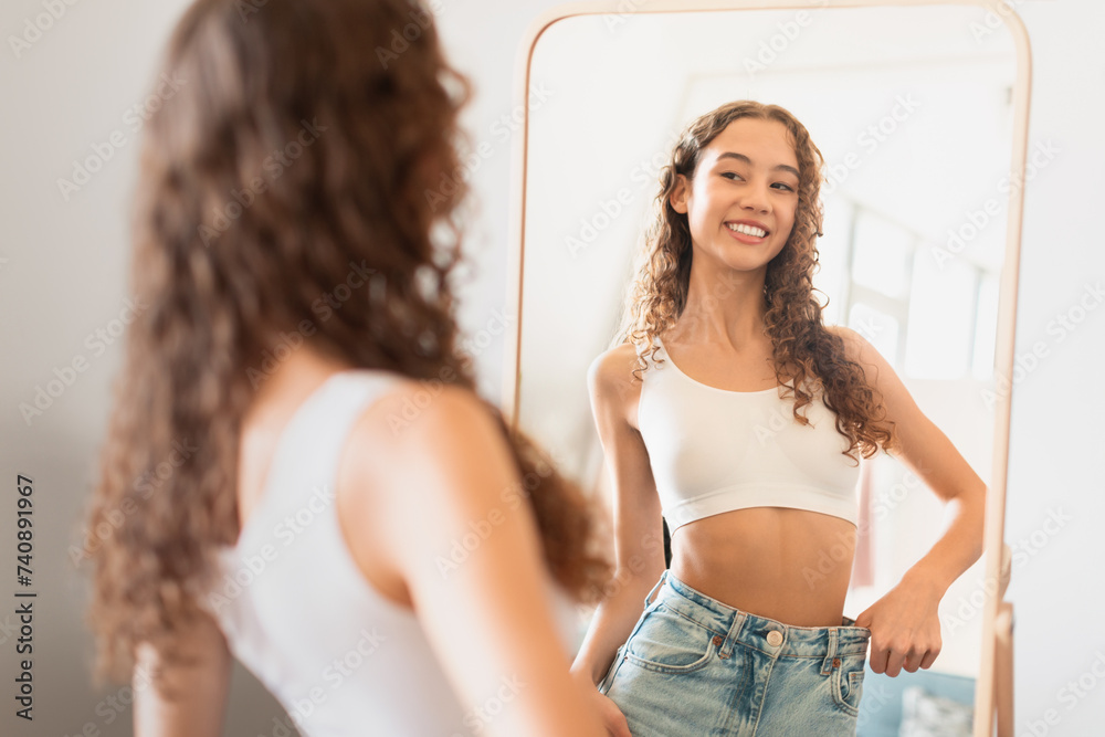Fototapeta premium teen girl looks at mirror satisfied with weight loss indoor