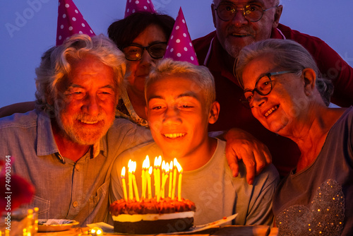 Grandfathers senior people and young teenager celebrate birthday together by night at home with cake and candles having fun and enjoying love and friendship at the party with different generations