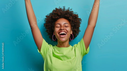 joyful person with curly hair, raising their arms in excitement, wearing a green shirt and red earrings, with a wide, laughing smile against a blue background.