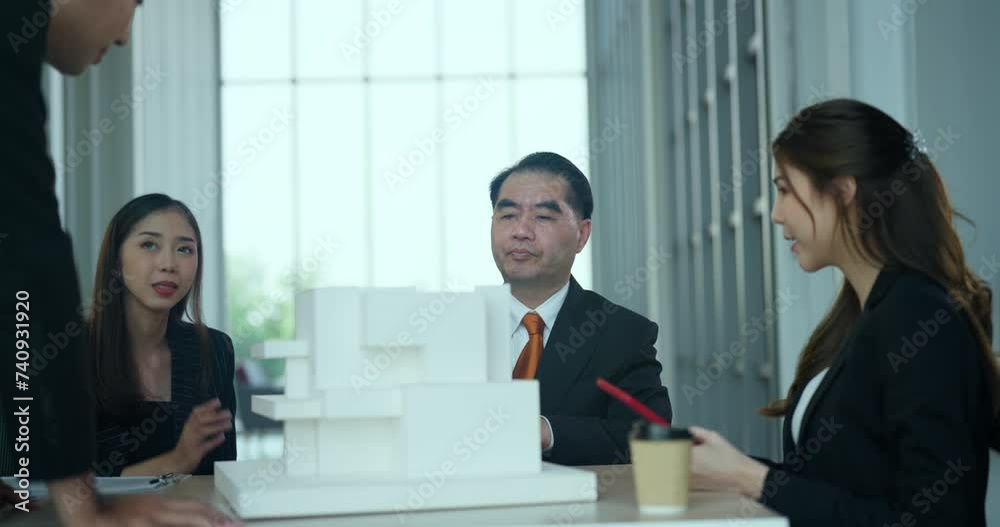 Group of business professionals gathers around a table, engaging in lively discussion while examining a detailed building model. Group of business professionals gathers around a table, engaging in lively discussion while examining a detailed building model.