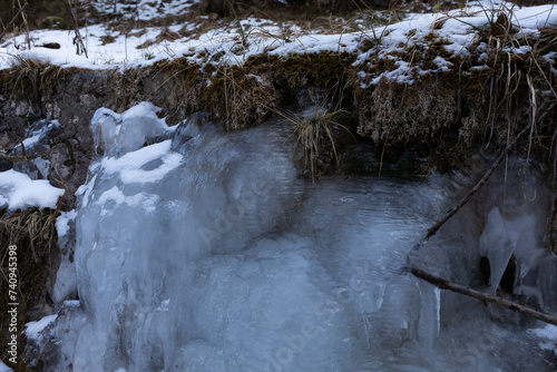 visuale in primo piano di una piccola cascata d'acqua completamente ghiacciata, lungo un leggero pendio erboso ed innevato in un bosco di montagna, in inverno photo