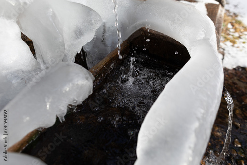 vista in primo piano di gocce d'acqua, che cadono all'interno di una grande bacinella in legno, da un rubinetto all'aperto, in montagna, coperto e circondato da ghiaccio e neve, in inverno photo