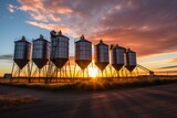 The sun sets behind a line of grain silos, casting long shadows and a radiant glow across the agricultural heartland