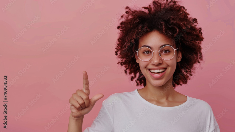 An exuberant young woman with curly hair and glasses, pointing her finger to the side with a wide, joyful smile on a pink background. An exuberant young woman with curly hair and glasses, pointing her finger to the side with a wide, joyful smile on a pink background.