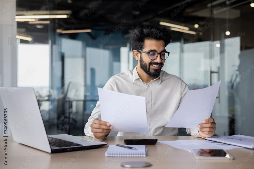 Satisfied Indian young man businessman working with documents, sitting in the office at the table and looking through the papers Satisfied Indian young man businessman working with documents, sitting in the office at the table and looking through the papers