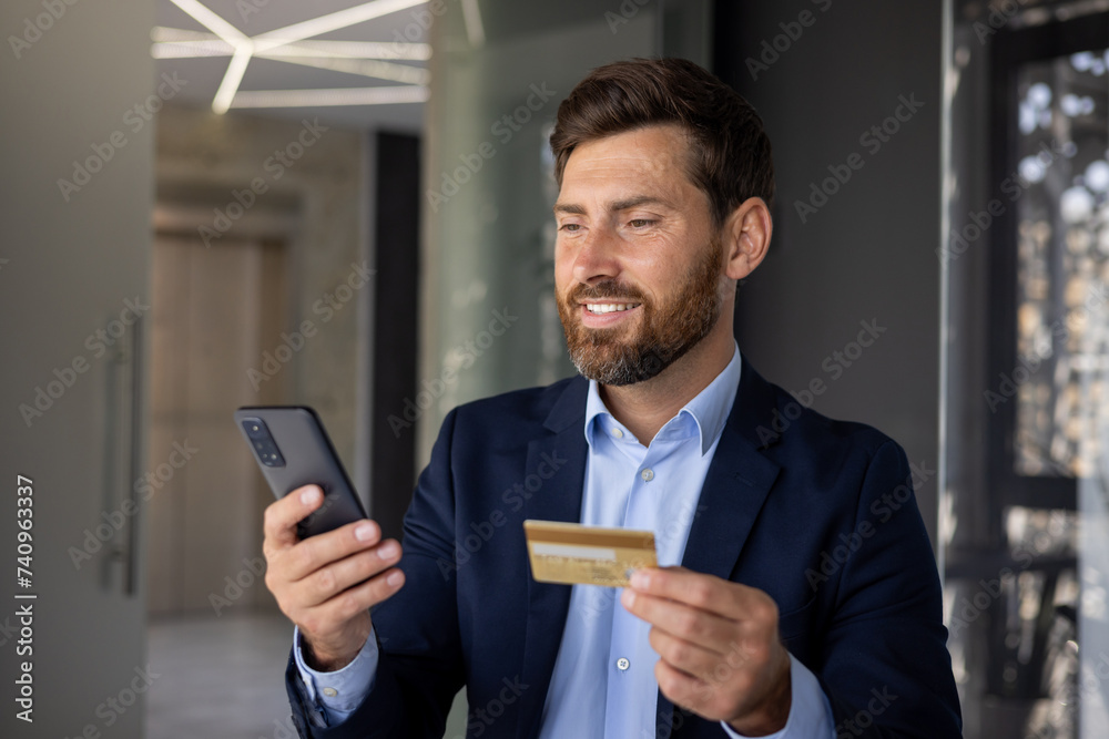 Close-up photo of a young businessman standing inside an office center in a suit, holding a credit card and using a mobile phone