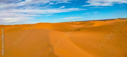 Fototapeta Naklejka Na Ścianę i Meble -  A sweeping panorama captures the vast expanse of golden sand dunes stretching as far as the eye can see, forming an arid desert under a partly cloudy blue sky in Timimoun, Algeria.