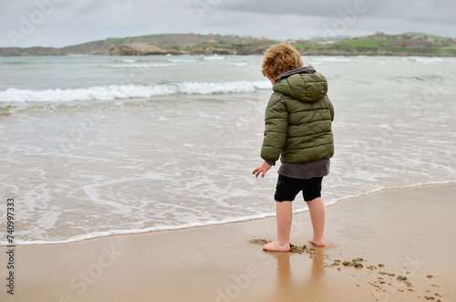 Portrait of a little kid on his back walking barefoot on the beach sand with his coat on on a cold winter day playing with the waves. Free play in childhood and connection with nature. Copy-space.