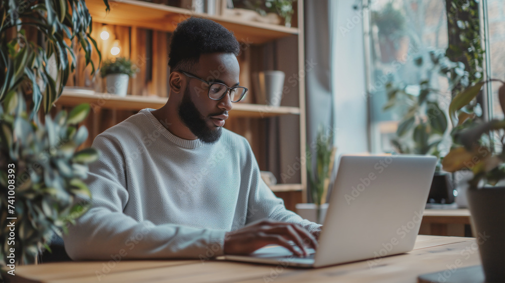 A diligent man with glasses focused intently on his laptop screen, in a cozy and plant-filled workspace, embodying the blend of nature and technology in modern work life