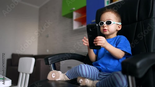 Sweet baby boy wearing sunglasses sits in office chair. Kid looks at smartphone in his hands. Low angle view. photo