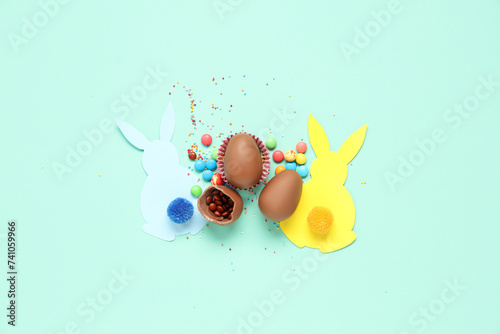 Sweet chocolate Easter eggs with candies and bunnies made of paper on turquoise background