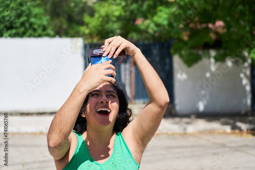 Smiling young latin woman watching an eclipse of the sun with eclipse glasses, taking photo with phone