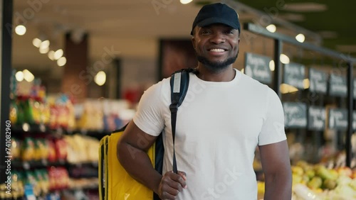 Portrait of a happy male delivery worker with Black skin color in a black hat and a white T-shirt who carries a large yellow bag on his shoulders while searching for the necessary goods in a grocery photo
