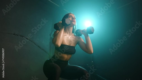 In a moody gym setting, a woman combines a squat with bicep curls, showcasing her multi-tasking ability in fitness as she works on both strength and technique under subtle lighting. 