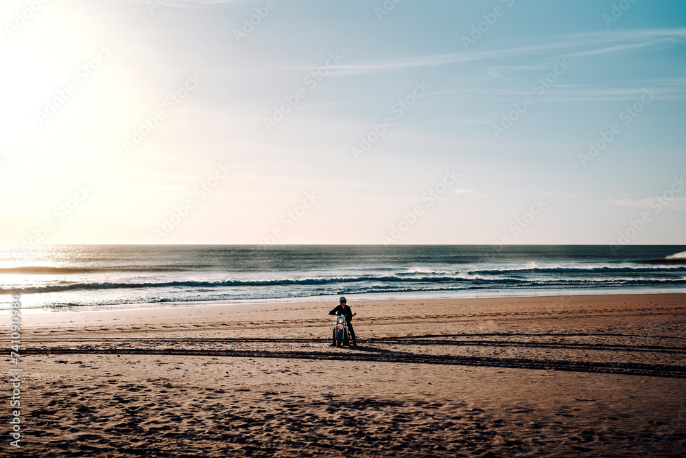 Chico parado en mitad de la playa con su moto de campo al atardecer