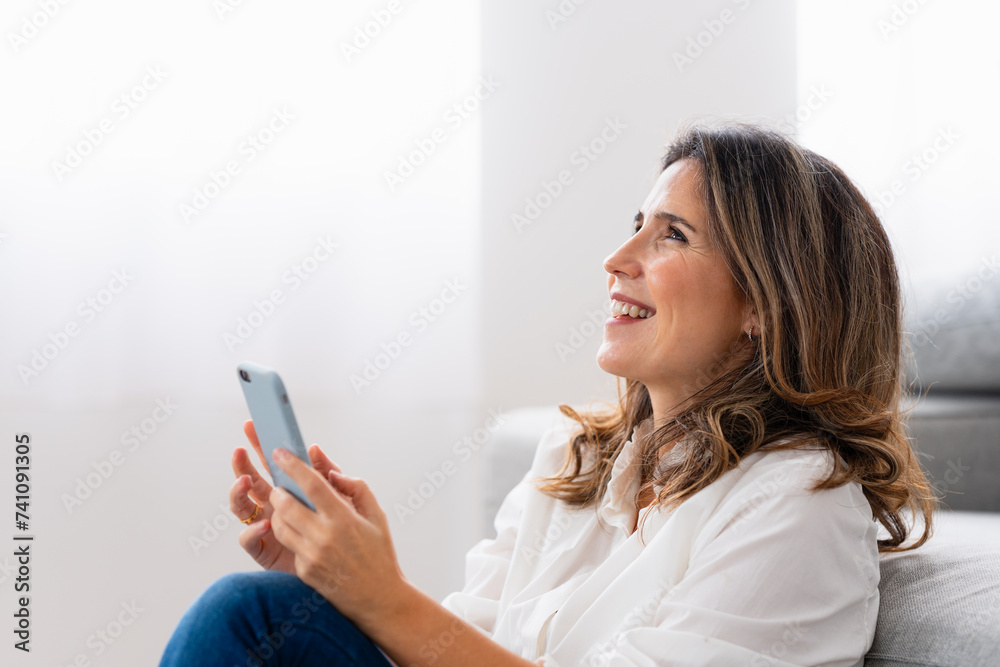 Woman smiling while using phone sitting on the floor in the living room Woman smiling while using phone sitting on the floor in the living room