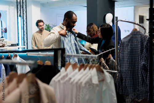 Boutique client trying on casual plaid shirt, examining size and quality while shopping with assistant. Clothing store consultant holding apparel to customer body and giving fashion advice