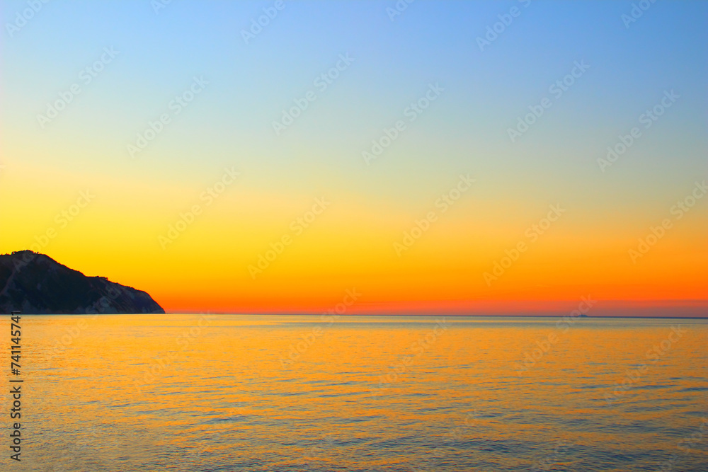 Astonishing sunset view from Le Terrazze di Portonovo, a sky perfectly imbued with gradient tones from blue to orange to red, the calm golden surface of the Adriatic Sea, and a mountain on the horizon