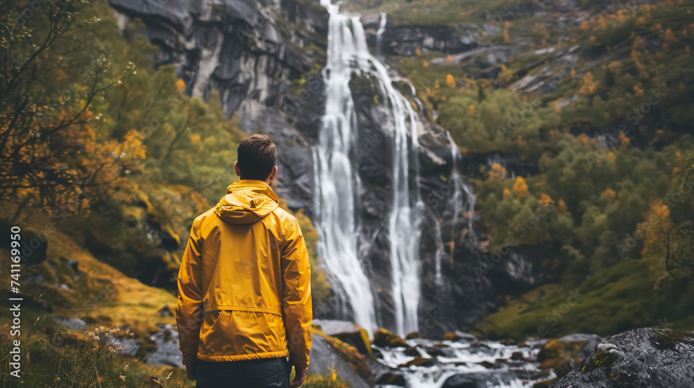 A back view of man wearing a yellow hoodie jacket on waterfall nature background.