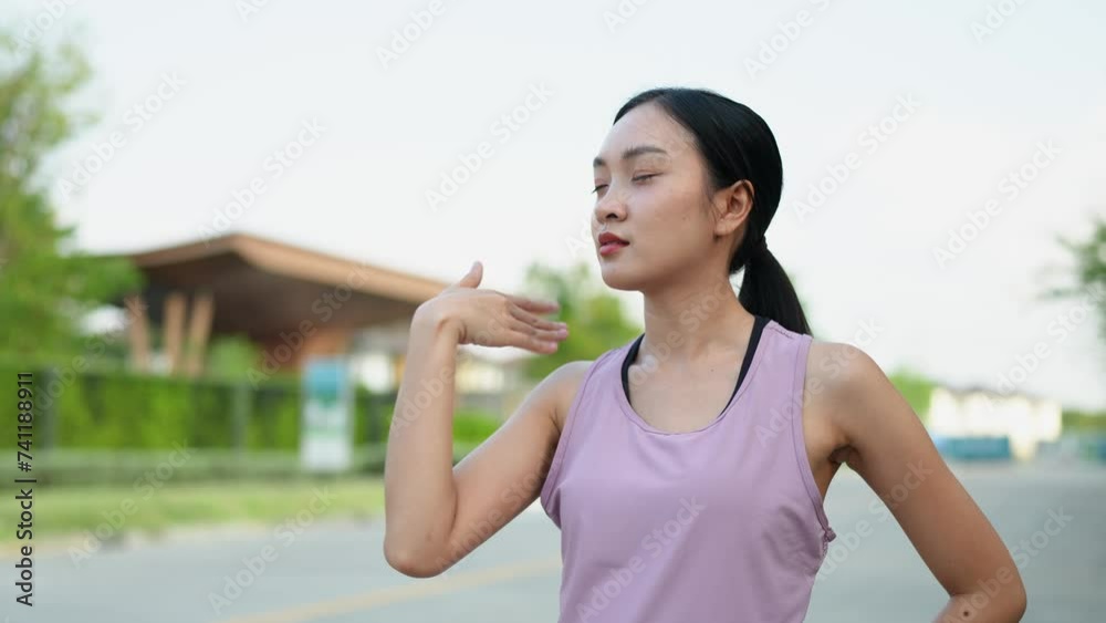 Enthusiastic Asian female runner stands using a hand fan to relieve heat and shortness of breath after running in the park. Female athlete takes a break after morning jogging. Enthusiastic Asian female runner stands using a hand fan to relieve heat and shortness of breath after running in the park. Female athlete takes a break after morning jogging.
