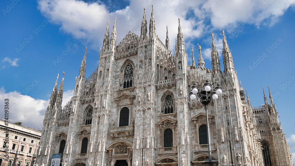 The intricate façade of the Milan Cathedral, with its numerous spires and sculptures, against a backdrop of white clouds and blue sky.