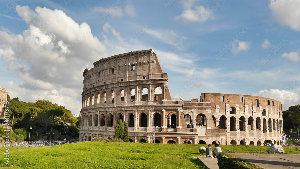 The Colosseum stands under a sunny sky, surrounded by greenery and ruins, showcasing its iconic ancient Roman architecture.