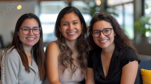 Women empower and support each other in the workplace. Diversity of nationality, Latino and Asian women. Three employee are sitting and smiling beside each other on a blurred workplace background.