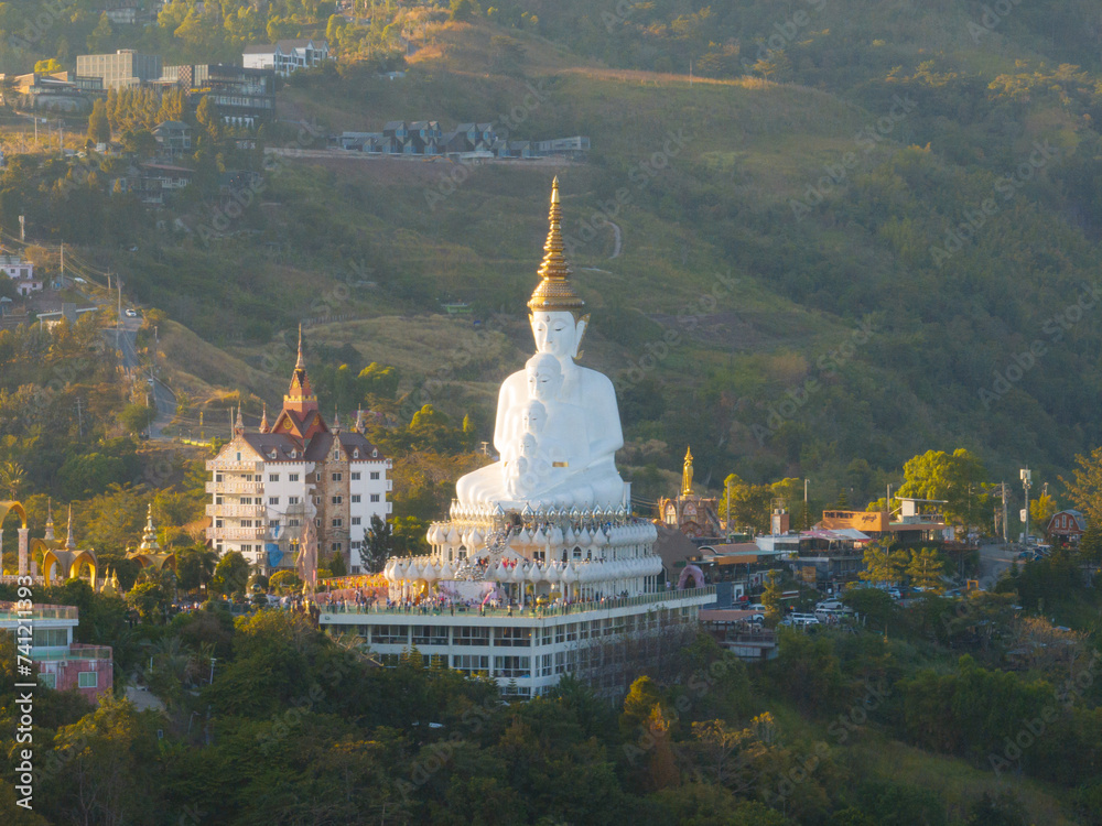 Aerial view of a majestic white Buddha statue atop a hill with a scenic sunset backdrop, overlooking a bustling town below.